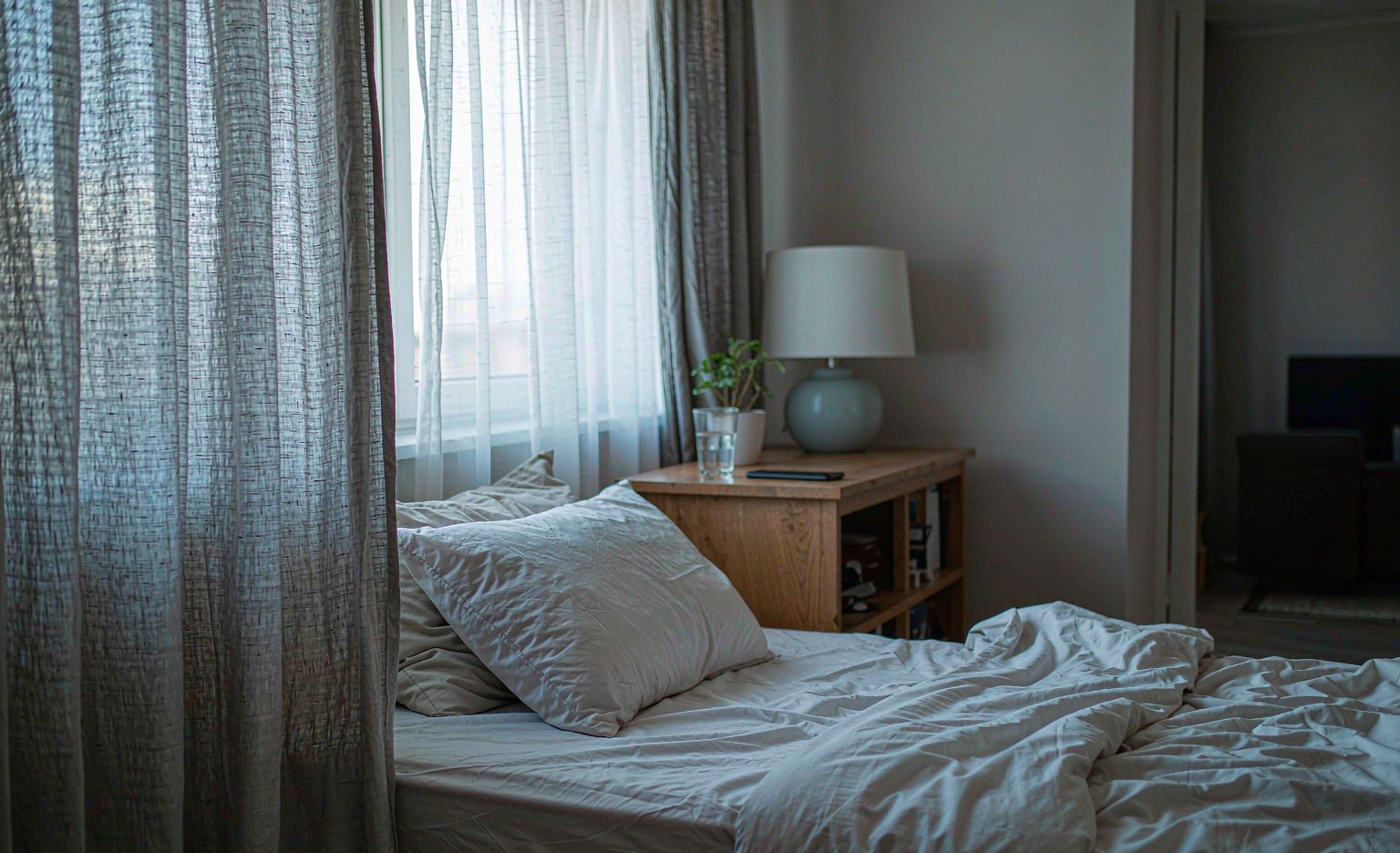 A bedroom at dawn with an unmade bed, a phone left face-down on the nightstand and pale light through a linen curtain