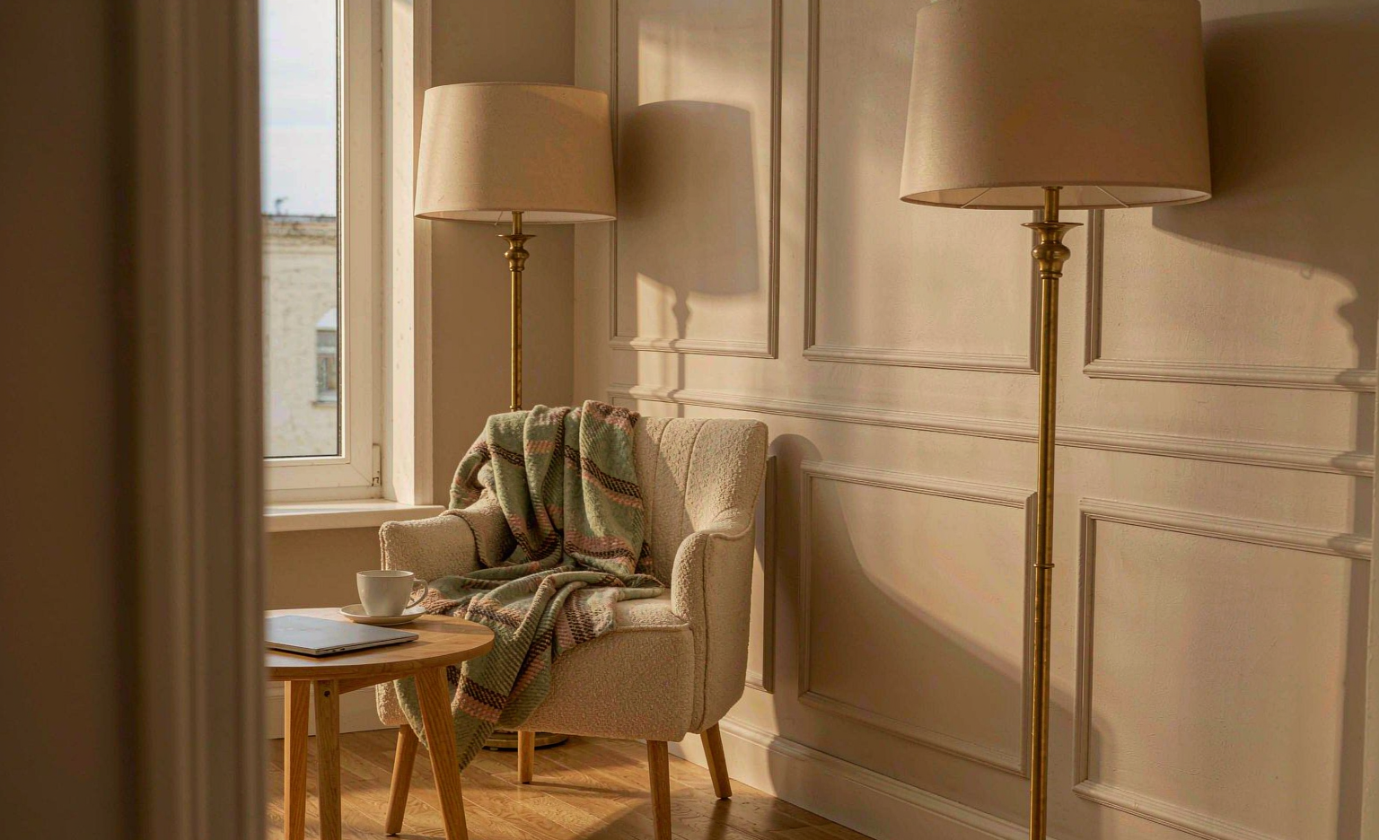 A quiet corner of a room with an upholstered lounge chair, a folded blanket, a closed laptop and a cup on a side table in late afternoon light