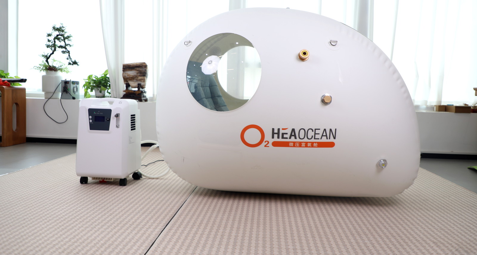 A white HEAOCEAN sit-and-lie chamber in a showroom, with its observation window, brass ports and zip visible, and an oxygen concentrator alongside